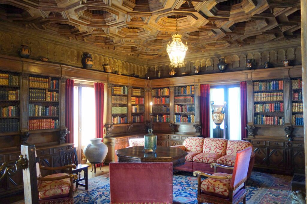 Interior view of the Library at Hearst Castle, featuring carved wooden ceilings, classical columns, and shelves lined with antique books and decorative art objects, reflecting William Randolph Hearst’s eclectic collecting style.